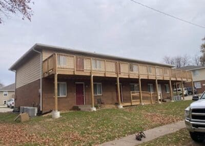 Long wooden deck and walkway with railing attached to a two-story brick and siding building, surrounded by grass and a sidewalk, illustrating custom outdoor construction for decks, walkways, and ramps.