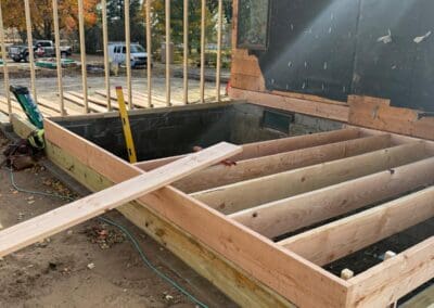 Construction of a wooden deck frame with evenly spaced joists, built on a foundation near a building wall, showcasing early-stage outdoor deck craftsmanship focused on durability and custom design.